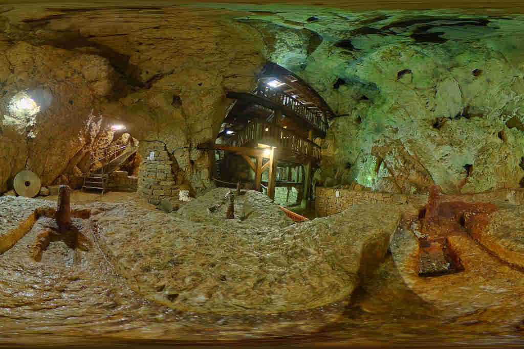 Photo des Moulins souterrains du Col-des-Roches, canton de Neuchâtel, Suisse. Grotte avec éclairage artificiel très lumineux avec reflets verts. Escalier descendant à gauche, structure en bois à droite sur plusieurs étages
